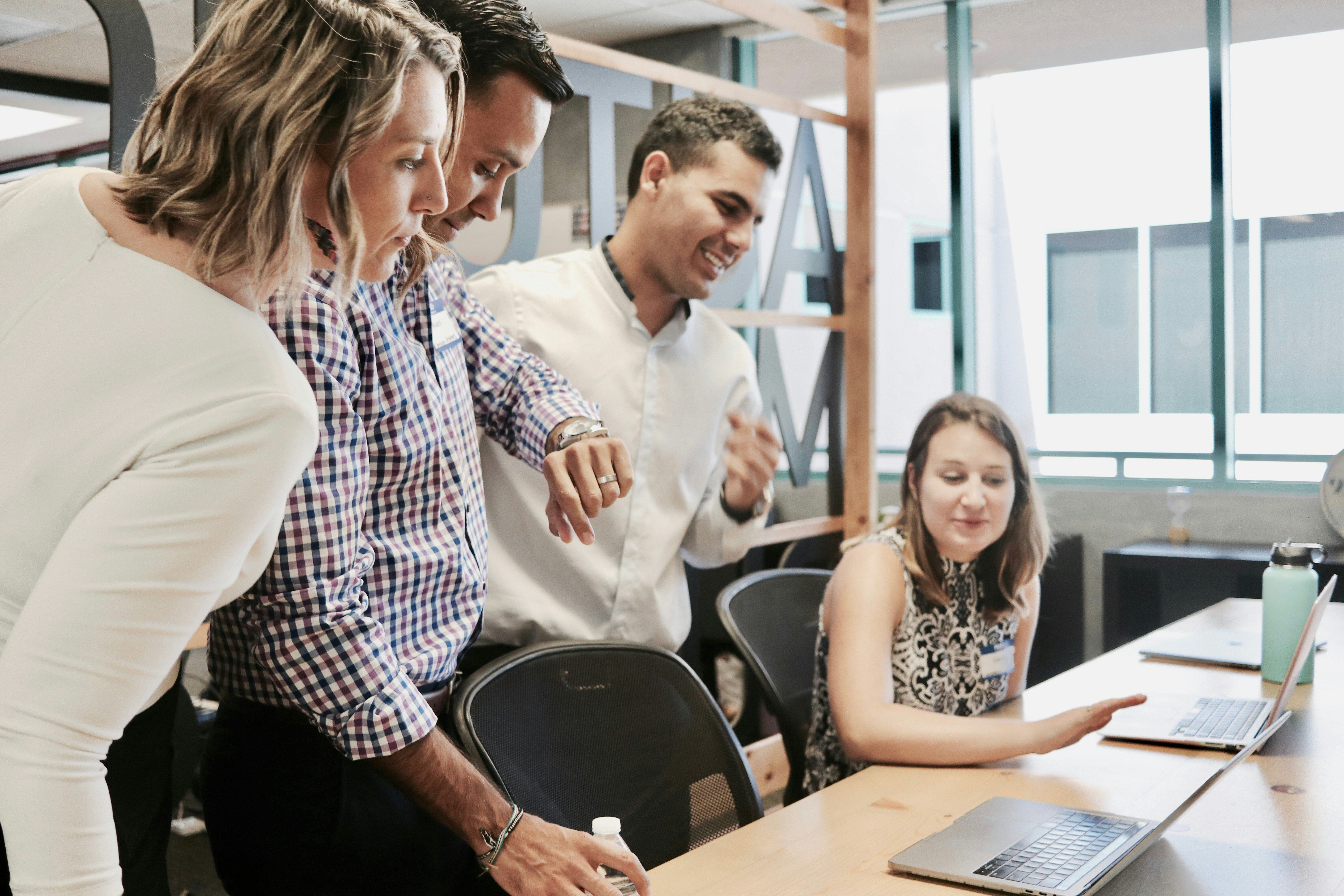 Two men and two women look at a laptop on a desk, one man looking at his watch