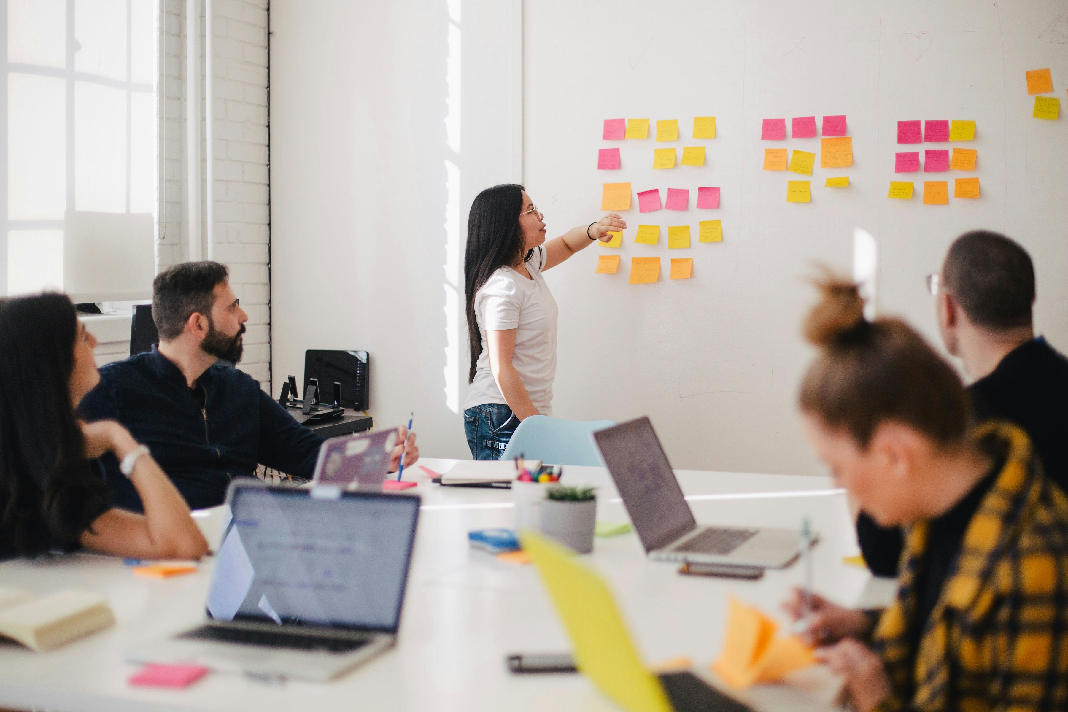Woman standing in front of a whiteboard with sticky notes, while coworkers with laptops sit at a table